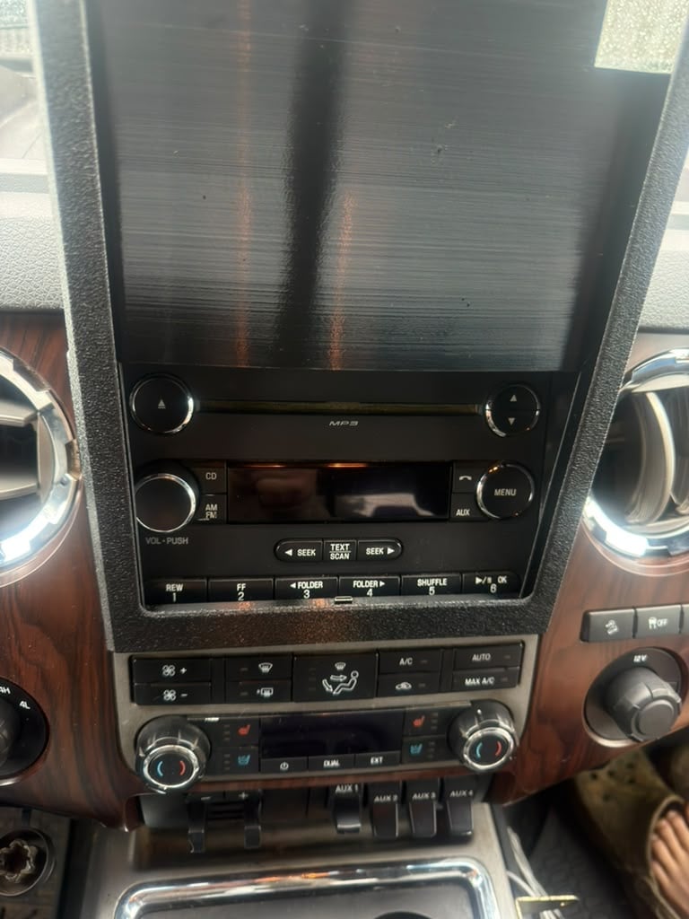 Vertical car dashboard touchscreen display with chrome bezels, climate controls, and knobs below mounted in dark wood trim