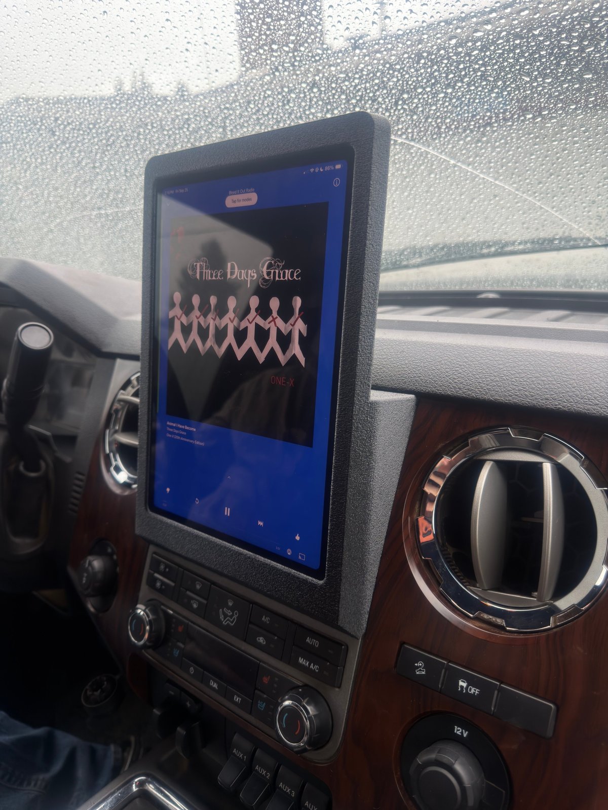 Touchscreen infotainment display in a car showing music player with album artwork on rainy windshield