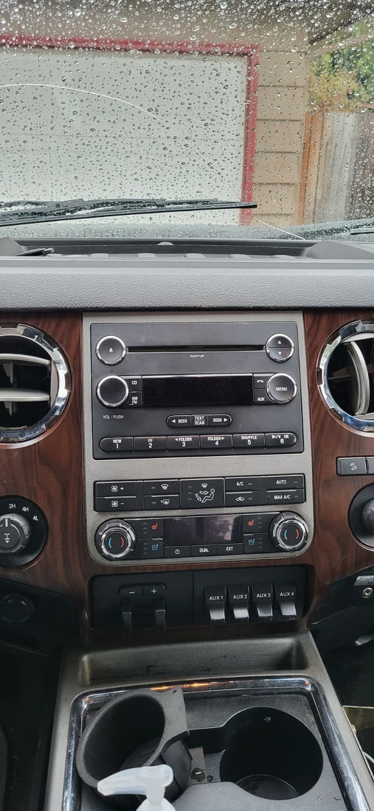 Car interior dashboard with wood trim, climate controls, and cup holders viewed from rainy windshield